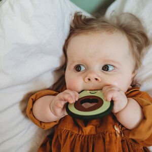 Puede incluir: Un beb&eacute; con un vestido marr&oacute;n est&aacute; mordiendo un mordedor verde en forma de aguacate con un agujero en forma de coraz&oacute;n y una cara sonriente.