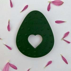 Puede incluir: Un mordedor de silicona verde oscuro con forma de aguacate con un corte en forma de coraz&oacute;n y el texto "Little Teethers" y "HEXNEX" en &eacute;l.