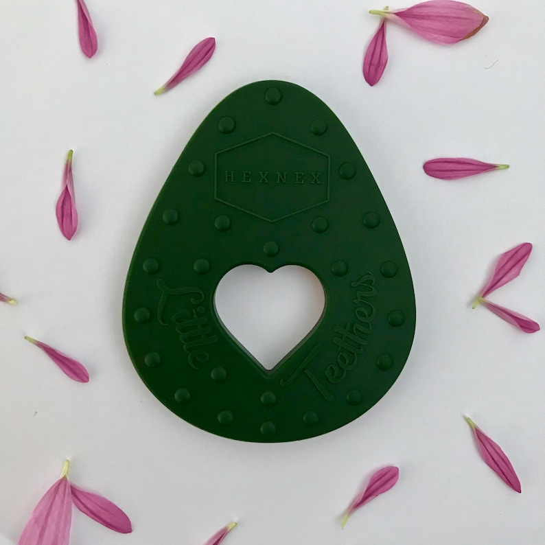 Puede incluir: Un mordedor de silicona verde oscuro con forma de aguacate con un corte en forma de coraz&oacute;n y el texto "Little Teethers" y "HEXNEX" en &eacute;l.