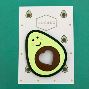 Puede incluir: Un mordedor de silicona en forma de aguacate verde con un hueso marr&oacute;n y un recorte en forma de coraz&oacute;n. El mordedor tiene una cara sonriente con mejillas rosadas y ojos negros.