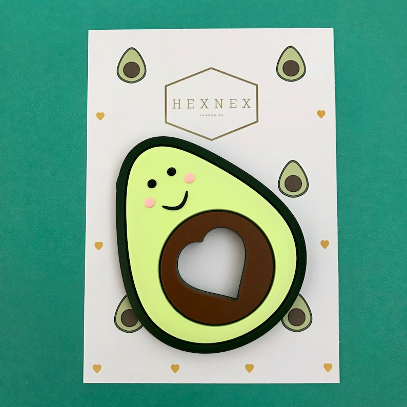 Puede incluir: Un mordedor de silicona en forma de aguacate verde con un hueso marr&oacute;n y un recorte en forma de coraz&oacute;n. El mordedor tiene una cara sonriente con mejillas rosadas y ojos negros.