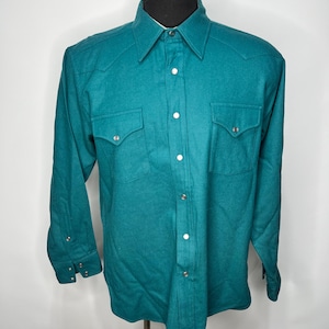 May include: Teal-colored button-down shirt with two chest pockets and silver-tone buttons. The long-sleeved shirt has a classic collar and is displayed on a mannequin. The shirt is made of a soft, possibly wool, material.
