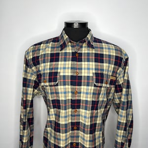 May include: A long-sleeved plaid shirt in a classic pattern. The shirt features a button-down front, two chest pockets, and a collar. The plaid design incorporates navy blue, red, and light blue lines on a cream background. The shirt is displayed on a mannequin.