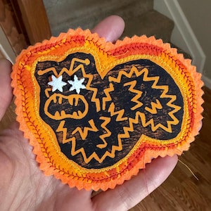 May include: A fabric patch featuring a black center with an orange zigzag border and orange zigzag line details. The patch includes two small white star-shaped accents positioned as eyes on the left side, with a jagged, irregular overall shape.