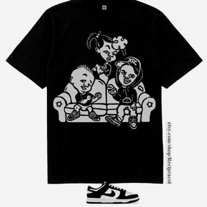 May include: Black and white graphic print t-shirt featuring three cartoon characters sitting on a couch. The characters are wearing casual clothing and have exaggerated features. The t-shirt is on a white background.