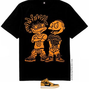 May include: Black t-shirt with an orange graphic of two cartoon characters, one with dreadlocks and glasses, the other with a shaved head. The characters are wearing streetwear and sneakers. The text "J.T.L." is visible on the character's shirt.  A pair of orange and black sneakers are also pictured.