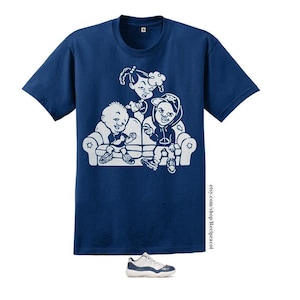 May include: A navy blue t-shirt with a white graphic of three cartoon characters sitting on a couch. The characters are wearing casual clothing and have a relaxed expression. The text "etsy.com/shop/Recipracol" is printed on the bottom right corner of the shirt. A pair of white and navy blue sneakers are also pictured.
