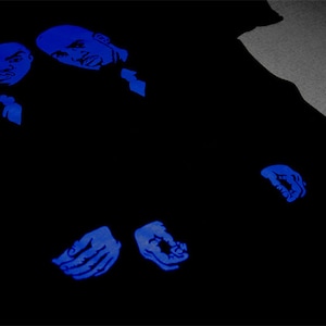 May include: A black t-shirt featuring a graphic print in vibrant blue ink. The design displays two stylized male faces positioned at the top left, with two hands shown near the bottom center, all rendered in a bold, monochromatic blue tone.