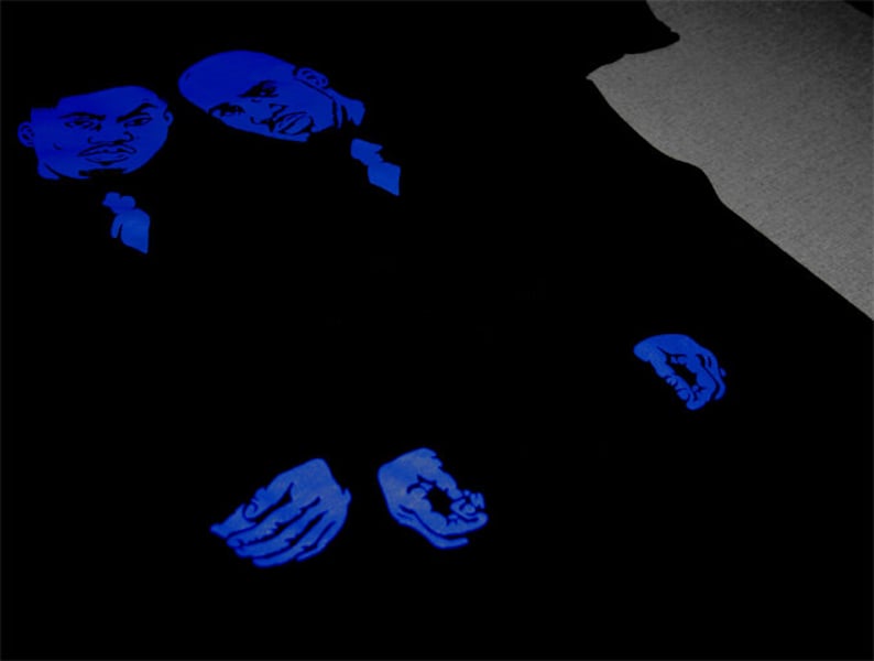 May include: A black t-shirt featuring a graphic print in vibrant blue ink. The design displays two stylized male faces positioned at the top left, with two hands shown near the bottom center, all rendered in a bold, monochromatic blue tone.