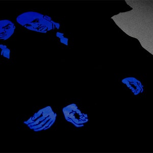 May include: A black t-shirt featuring a graphic print in vibrant blue ink. The design displays two stylized male faces positioned at the top left, with two hands shown near the bottom center, all rendered in a bold, monochromatic blue tone.