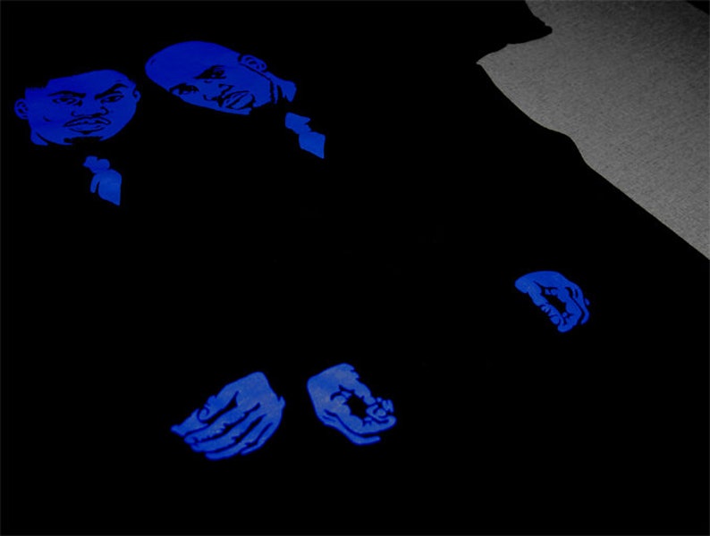 May include: A black t-shirt featuring a graphic print in vibrant blue ink. The design displays two stylized male faces positioned at the top left, with two hands shown near the bottom center, all rendered in a bold, monochromatic blue tone.