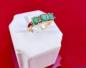 May include: A gold ring with three oval-shaped emerald gemstones and smaller diamond accents. The ring is displayed on a white box with a red background.