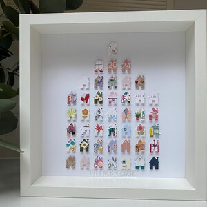 May include: A square white shadow box frame containing a collection of small, house-shaped paper cutouts arranged in a triangular formation. The cutouts feature various floral, geometric, and solid patterns. The text 20 24 and THYME CLOSE are visible.
