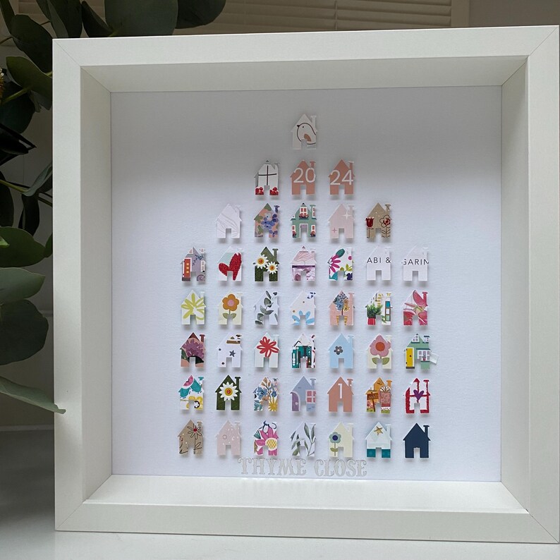 May include: A square white shadow box frame containing a collection of small, house-shaped paper cutouts arranged in a triangular formation. The cutouts feature various floral, geometric, and solid patterns. The text 20 24 and THYME CLOSE are visible.