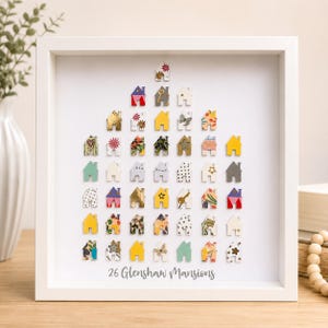 May include: A white square shadow box frame containing a collection of small, multi-coloured house-shaped cutouts arranged in a triangular pattern. The text 26 Glenshaw Mansions is printed in grey at the bottom centre of the white background.