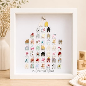 May include: A white framed wall art piece featuring a collection of small, multi-coloured house-shaped cutouts arranged in a triangular shape. The cutouts display various patterns, colours, and the text "NEW HOME". The text "15 Oakwood Drive" is printed at the bottom.
