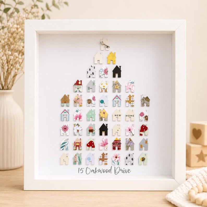 May include: A white framed wall art piece featuring a collection of small, multi-coloured house-shaped cutouts arranged in a triangular shape. The cutouts display various patterns, colours, and the text "NEW HOME". The text "15 Oakwood Drive" is printed at the bottom.