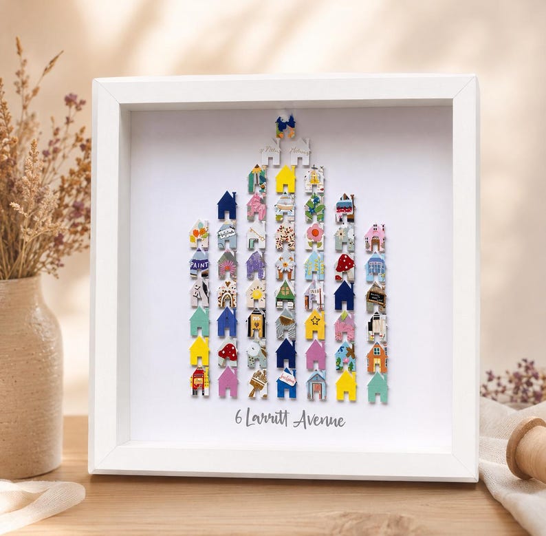 May include: A white square shadow box frame containing a collection of small, colourful house-shaped cutouts arranged in a tall, pointed formation. The text 6 Larritt Avenue is printed in grey at the bottom centre of the white backing board.