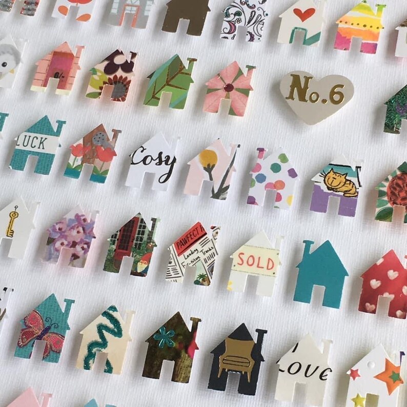 May include: A collection of small, die-cut paper house shapes featuring diverse patterns, colours, and illustrations. Some houses display text including 'LUCK', 'Cosy', 'PAWFECT', 'SOLD', and 'love', alongside a heart-shaped piece labelled 'No.6'.