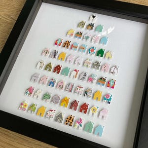 May include: A framed wall art piece featuring a collection of small, house-shaped cutouts arranged in a triangular tree formation. Each individual house cutout displays a unique pattern, colour, or graphic design, including floral, geometric, and text prints.