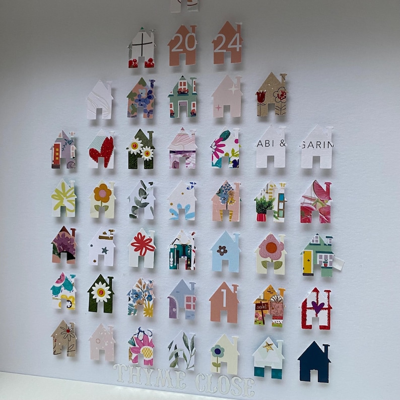 May include: A collection of small house-shaped paper cutouts arranged in a grid on a white surface. The houses feature various colourful patterns, floral prints, and geometric designs. Some houses display the text 2024, ABI & GARIN, and THYME CLOSE.