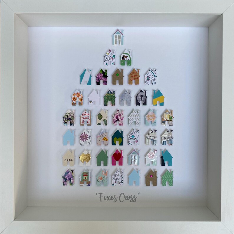 May include: A white framed wall art piece featuring a collection of small, house-shaped paper cutouts arranged in a triangular formation. Each house displays unique patterns, colours, and textures. The text 'Foxes Cross' is printed at the bottom.