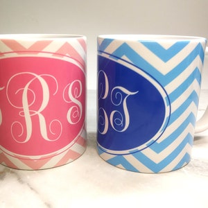 May include: Two ceramic mugs with chevron patterns. One mug is pink with a white monogram "TRS" and the other mug is blue with a white monogram "DT".