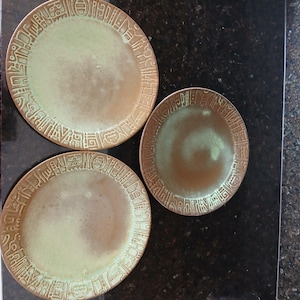 May include: Three round ceramic plates featuring a sage green center and a wide, textured border with an embossed geometric pattern in a bronze-brown tone. The plates have a smooth, glazed finish and a consistent design across all three pieces.