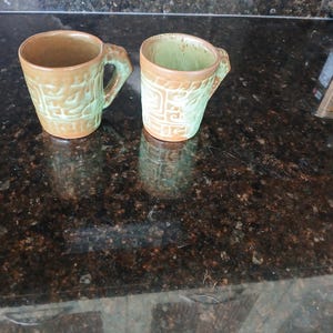 May include: Two ceramic mugs featuring a textured, embossed geometric pattern. The mugs have a two-tone glaze with a light green finish on the exterior and interior, accented by warm brown tones along the rims, bases, and handles.