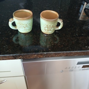 May include: Two ceramic mugs featuring a light green and tan glaze with a textured, embossed pattern on the exterior. Each mug has a rounded handle and a smooth, tan-colored interior rim, showcasing a rustic finish and a cylindrical shape.