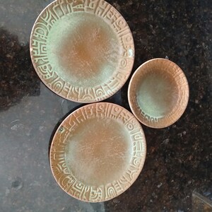 May include: Two round plates and one small bowl featuring a textured, metallic finish in shades of copper and verdigris green. Each piece displays a wide, embossed border decorated with repeating geometric patterns and abstract symbols.