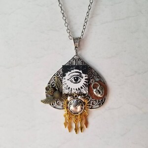Peut inclure: Un collier pendentif argenté avec un charme en forme de pique. Le charme présente un motif d'œil noir et blanc, un petit oiseau, un cercle couleur cuivre avec un cadenas et une horloge dorée avec des accents pendants.