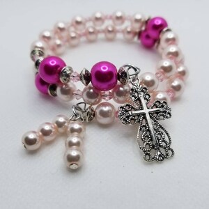K&ouml;nnte beinhalten: Ein doppelreihiges Armband mit rosa und wei&szlig;en Perlen und einem silbernen filigranen Kreuzanh&auml;nger. Das Armband hat einen Anh&auml;nger mit rosa Perlen.