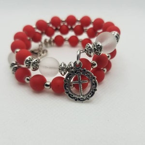 Può includere: Un braccialetto di perline rosse e bianche con un ciondolo d'argento a forma di croce. Il braccialetto è fatto di piccole perline rotonde e ha un design semplice ed elegante.