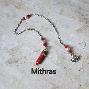 May include: A silver chain necklace with red and white beads and a red stone pendant. The pendant is a red, hexagonal stone with silver accents. The necklace also features a small silver charm and the word "Mithras".