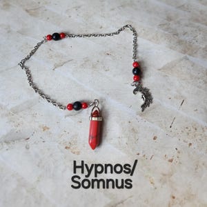 May include: A silver chain pendulum with a red and black beaded accent. The pendulum features a red crystal pendant and a silver crescent moon charm. The text "Hypnos/Somnus" is printed in black.