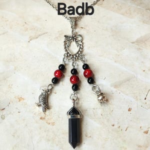 Peut inclure: Collier argenté avec un pendentif orné d'un cristal noir, de perles rouges et noires, et de breloques représentant un corbeau et un chaudron. Le mot "Badb" est écrit au-dessus.
