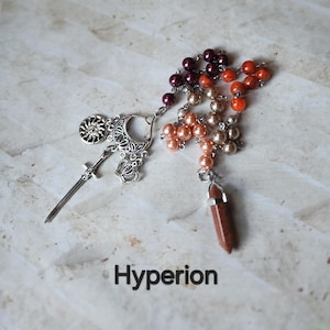 Puede incluir: Un collar artesanal con una espada, una corona y un dije de sol, con un degradado de cuentas de perlas en tonos burdeos, melocotón y naranja. Un colgante de cristal marrón cuelga en la parte inferior. La palabra "Hyperion" está impresa debajo.
