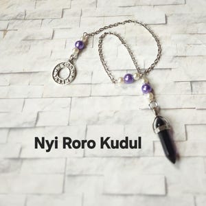 May include: A silver-toned metal chain pendulum featuring a dark purple crystal point, accented with small white and purple beads. The chain includes a circular metal charm with text and the words Nyi Roro Kudul printed below the object.