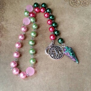 May include: A pink, green, and red beaded necklace with a silver charm of a tree with moons and a green and pink crystal pendulum.