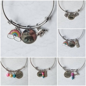 Puede incluir: Pulseras extensibles plateadas con varios dijes. Los dijes incluyen un arcoíris, un oso, las palabras "Mama Bear" y dijes en forma de corazón. Algunas pulseras tienen detalles de cuentas de colores. Las pulseras se exhiben sobre una superficie blanca.