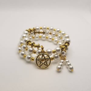 Puede incluir: Un brazalete de triple vuelta hecho con cuentas de perlas blancas y detalles dorados. El brazalete presenta un dije de pentáculo dorado y dos dijes de perlas colgantes.