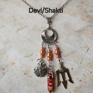 May include: Silver-toned necklace with a pendant featuring a crescent moon design, peach-colored beads, and dangling charms. Charms include a seashell, a red and yellow glass pendant, and a trident. The text "Devi/Shakti" is above the pendant.