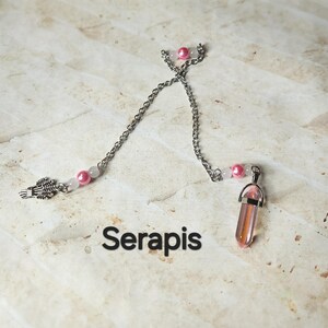 Puede incluir: Un collar de cadena plateada con cuentas rosas y blancas y un colgante de cristal rosa. El collar también presenta un dije en forma de espiga. La palabra "Serapis" está impresa en negro.