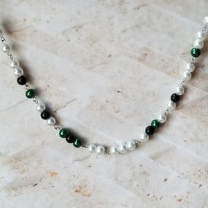 May include: A silver chain necklace with white, black, and green beads. The beads are arranged in a pattern.
