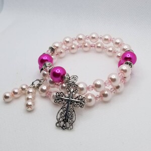K&ouml;nnte beinhalten: Ein doppelreihiges Armband mit rosa und wei&szlig;en Perlen und einem silbernen filigranen Kreuzanh&auml;nger.