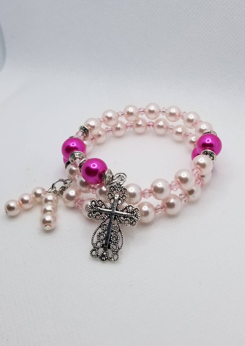 K&ouml;nnte beinhalten: Ein doppelreihiges Armband mit rosa und wei&szlig;en Perlen und einem silbernen filigranen Kreuzanh&auml;nger.