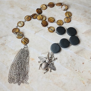 Op de afbeelding: Een ketting met ronde, schelpachtige kralen in bruin- en beigetinten, verbonden door een zilverkleurige ketting. Met een zilveren kwast en een bijenhangertje.