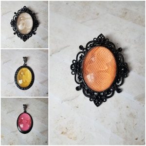 Può includere: Ciondoli e spille neri ornati con inserti ovali. Gli inserti presentano un motivo a pelle di serpente in vari colori, tra cui beige, giallo e rosa. Le spille hanno una cornice decorativa in stile filigrana.