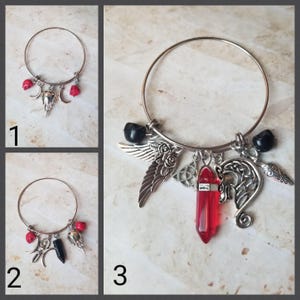 May include: Three silver bangle bracelets with different charms. The first bracelet has red beads, crescent moons, and a skull charm. The second bracelet has red beads, a crescent moon, a black spike, and a skull charm. The third bracelet has black beads, a silver wing charm, a red crystal charm, a Celtic knot charm, and a silver dragon charm.
